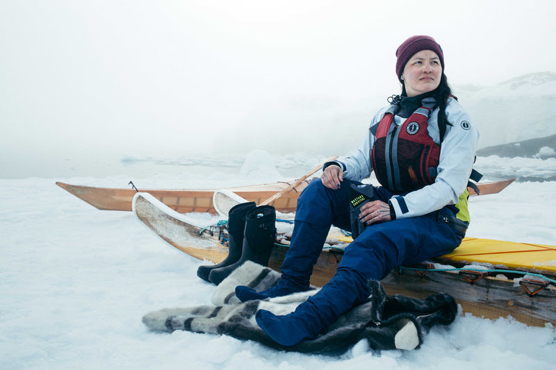 Amber sits on a wooden sledge in a snowy, icy landscape wearing a Destiny foam vest and a Helix drysuit.