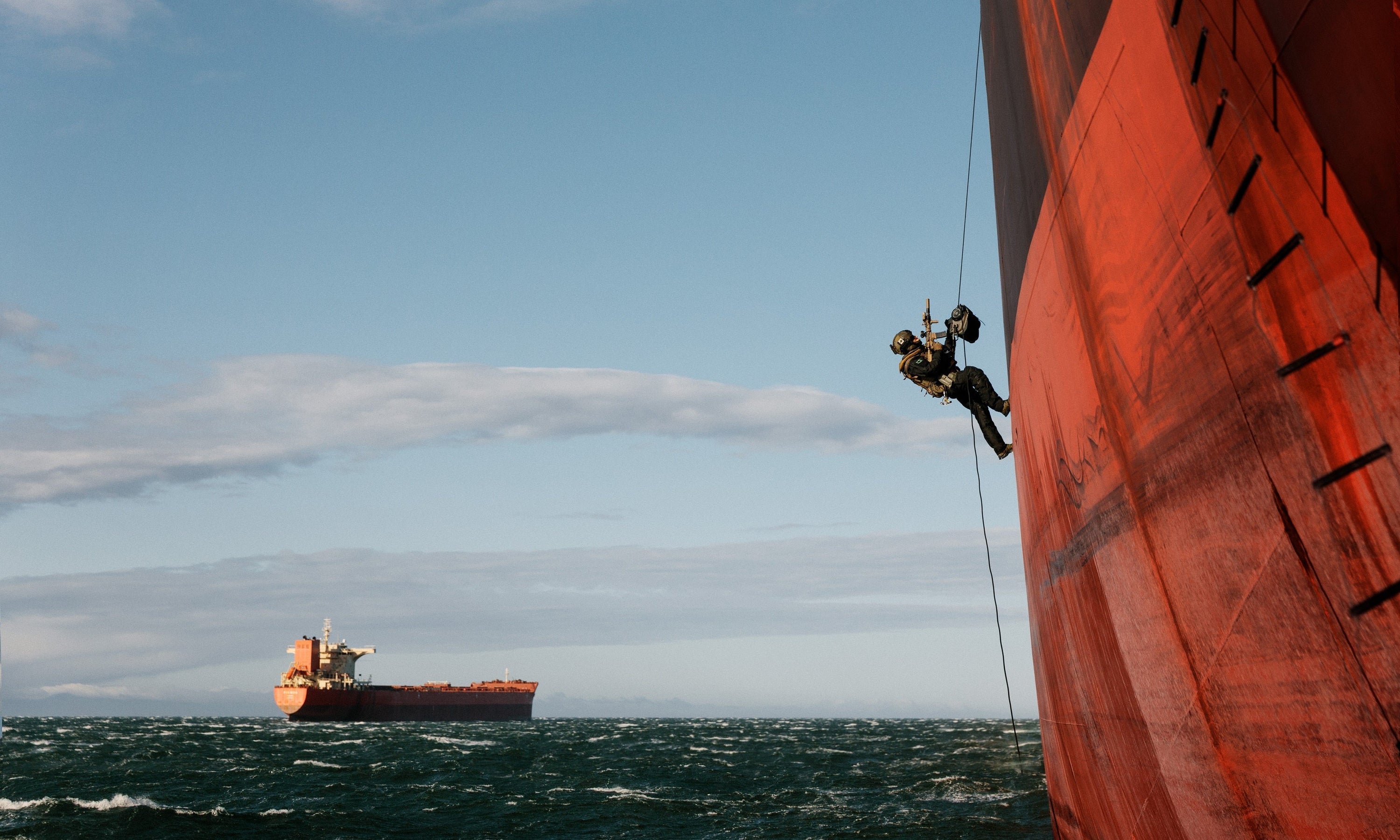 A tactical officer scales the massive red hull of a cargo ship on a rope over choppy seas.