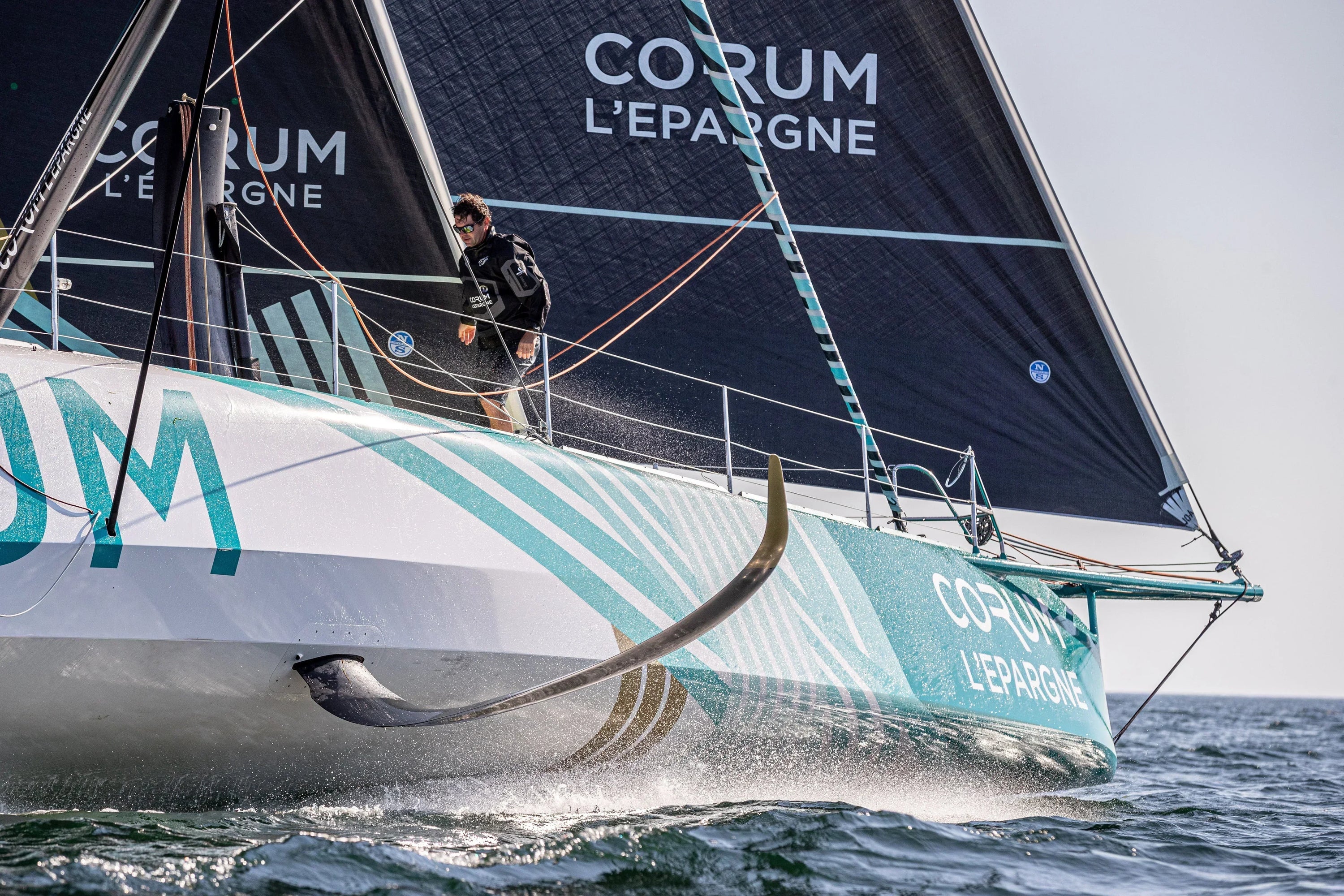 Skipper Nicolas Troussel stands on the deck of the CORUM L'Épargne IMOCA sailboat as it slices through the ocean.