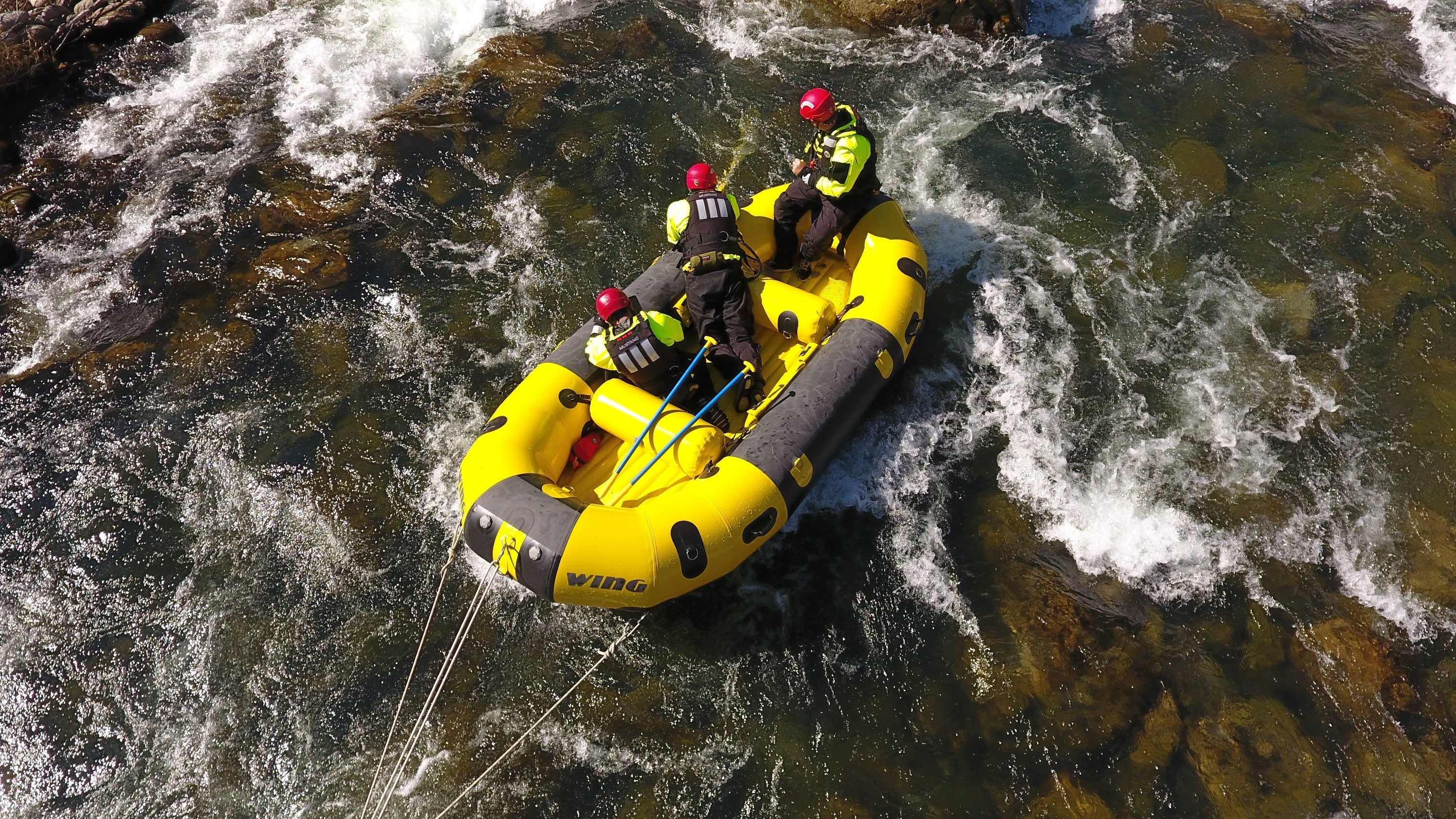 An aerial view of file 61edde.jpg shows three CAL FIRE rescue personnel wearing red helmets and high-visibility gear navigating a yellow inflatable raft through turbulent river rapids.
