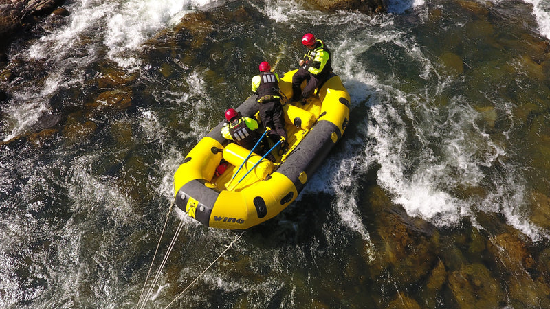 An aerial view of file 61edde.jpg shows three CAL FIRE rescue personnel wearing red helmets and high-visibility gear navigating a yellow inflatable raft through turbulent river rapids.