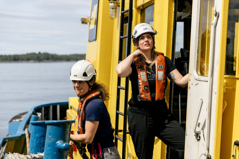 Hailey pulls a heavy mooring line while Ocean adjusts her safety helmet outside a bright yellow tugboat cabin.