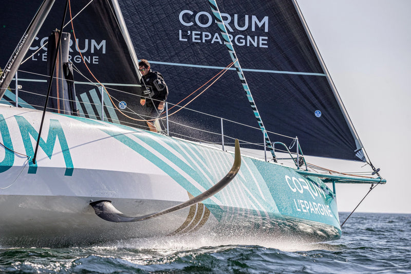 Skipper Nicolas Troussel stands on the deck of the CORUM L'Épargne IMOCA sailboat as it slices through the ocean.