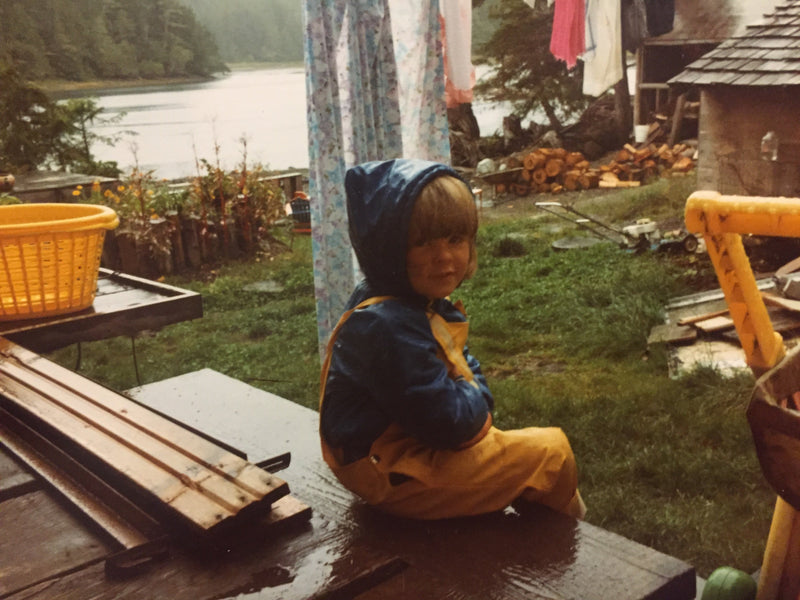 A vintage photo of young Danielle Baker in a blue and yellow rain suit, sitting on a wet porch in Bamfield, BC.