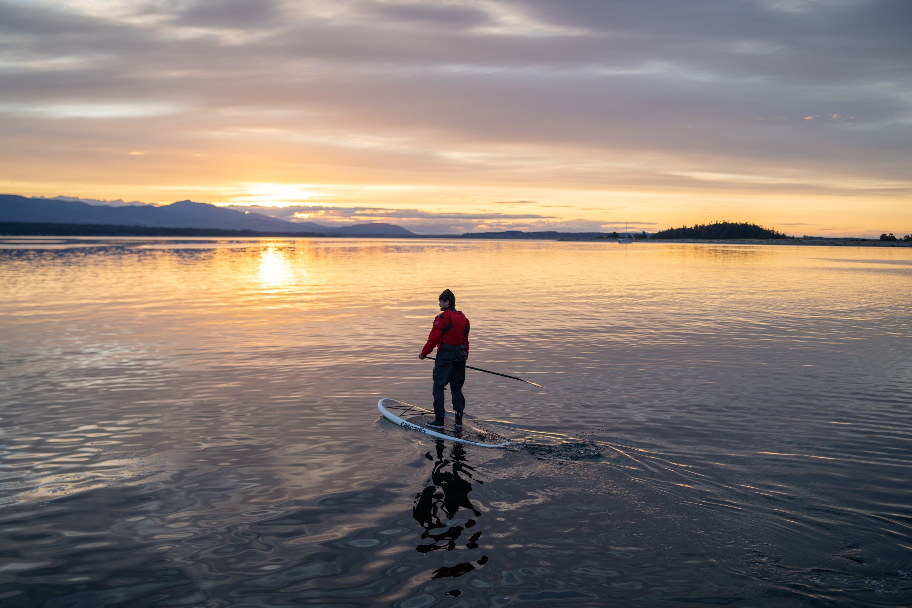 A person paddles a stand-up paddleboard on calm water during a golden sunset, with mountains silhouetted in the distance.