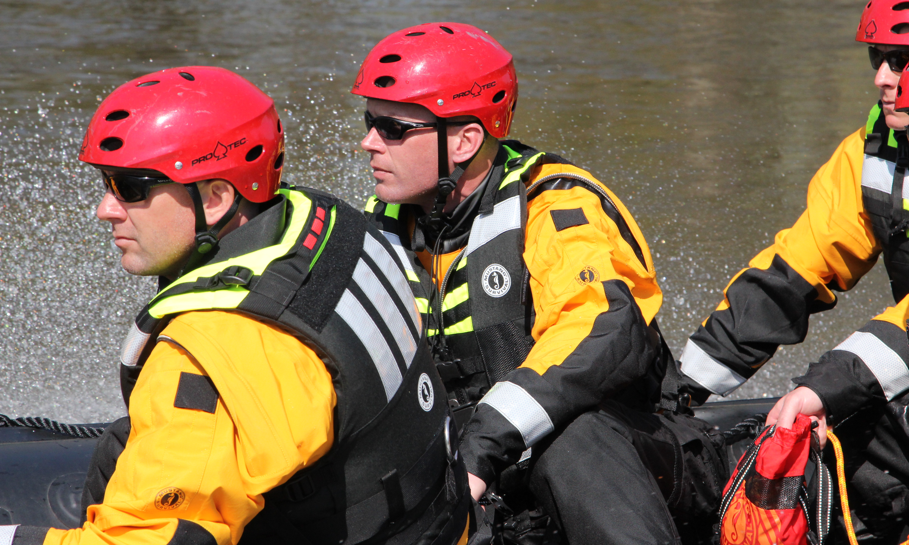 Three rescue personnel in red helmets and yellow dry suits ride in a boat; the foreground rescuer wears a Mustang Universal Swift Water Rescue Vest.
