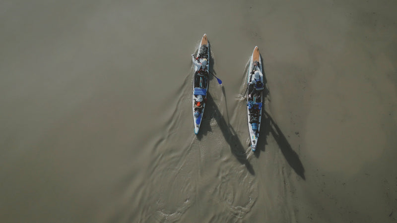 Overhead drone view of two people paddling expedition stand-up paddleboards side-by-side through murky river water.