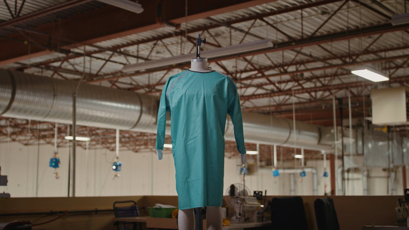 A teal medical gown hangs on a mannequin inside a large, industrial manufacturing facility with high exposed ceilings.