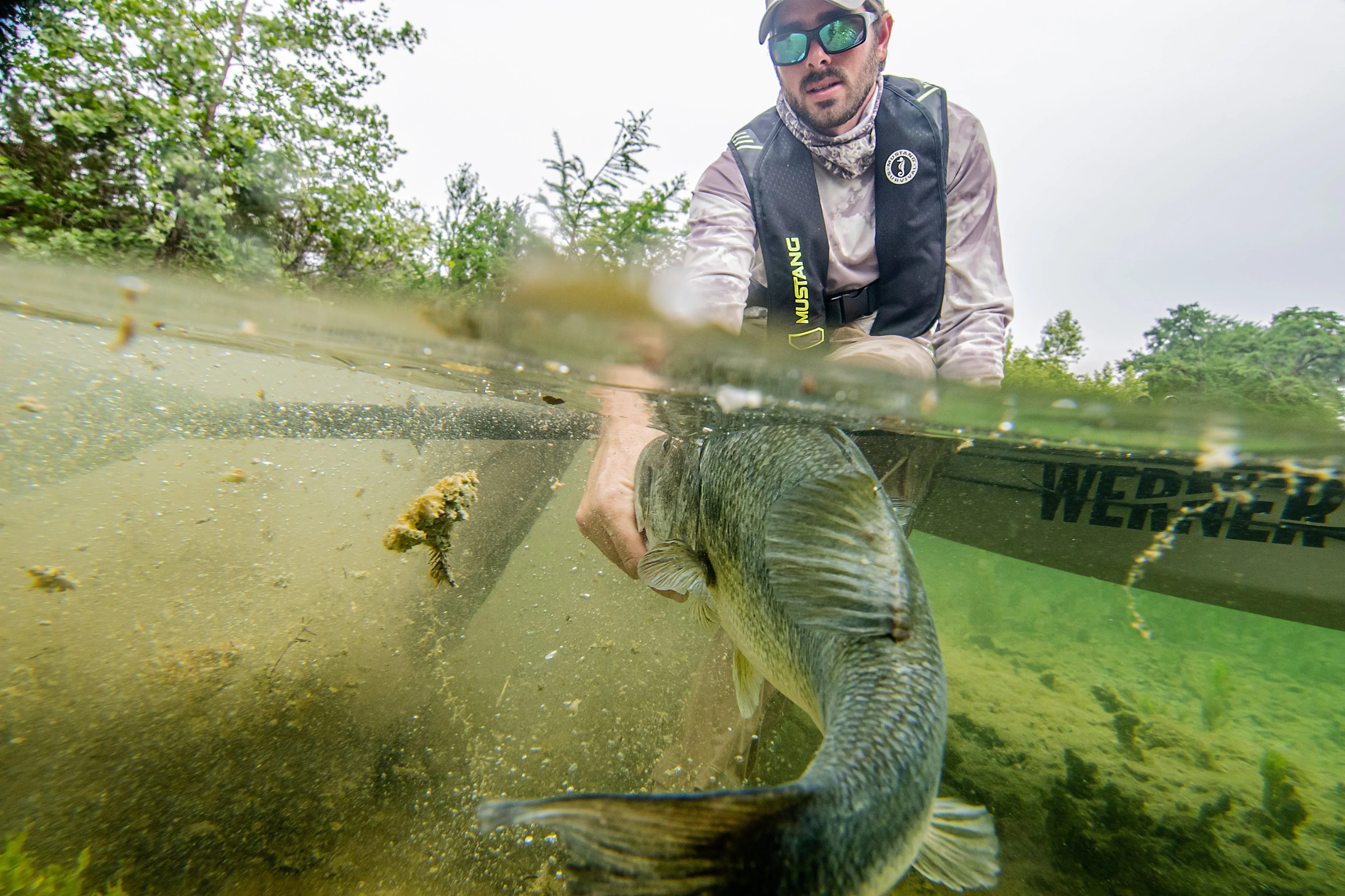 Man wearing a Mustang Survival life jacket, getting a bass into the water during a freshwater bass fishing trip, captured in a half-above, half-below water perspective.