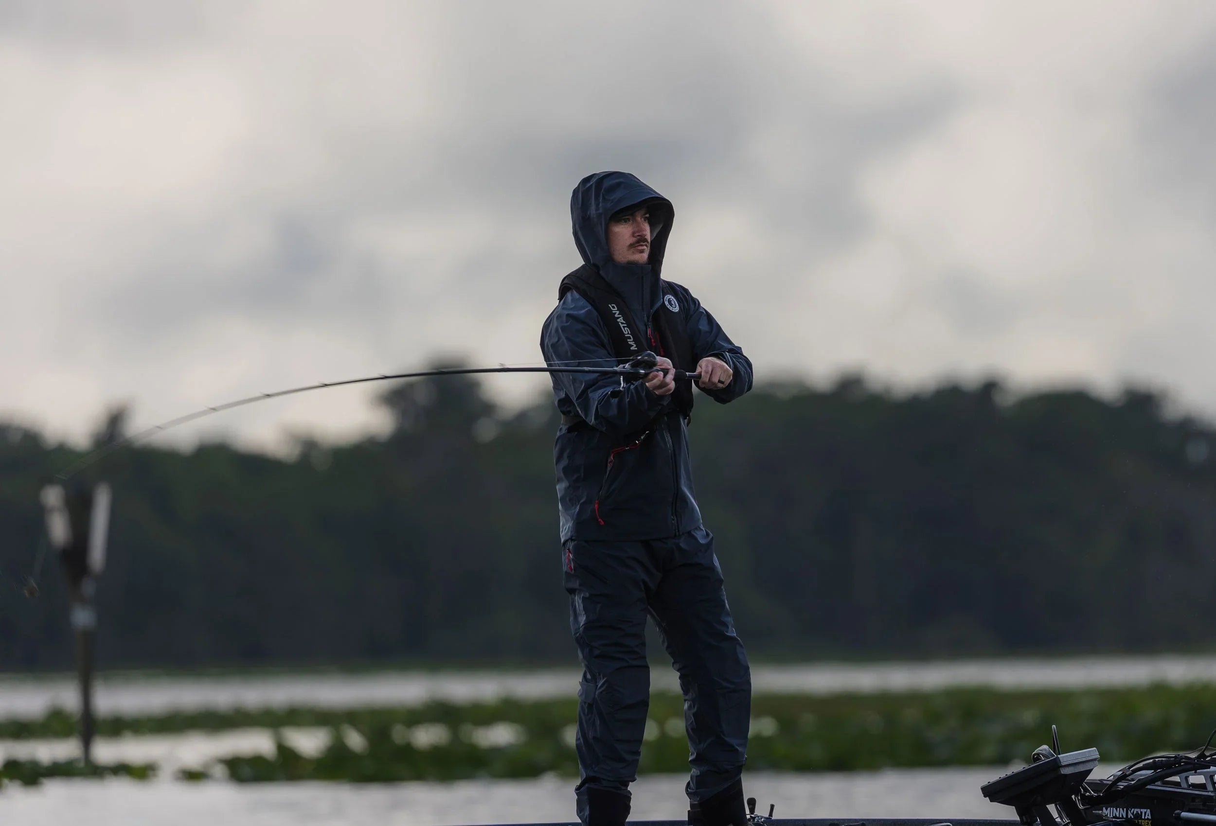 Person wearing a Mustang Survival jacket.d life jacket with hood, fishing on a lake from a boat in overcast weather, with forested shoreline in the background