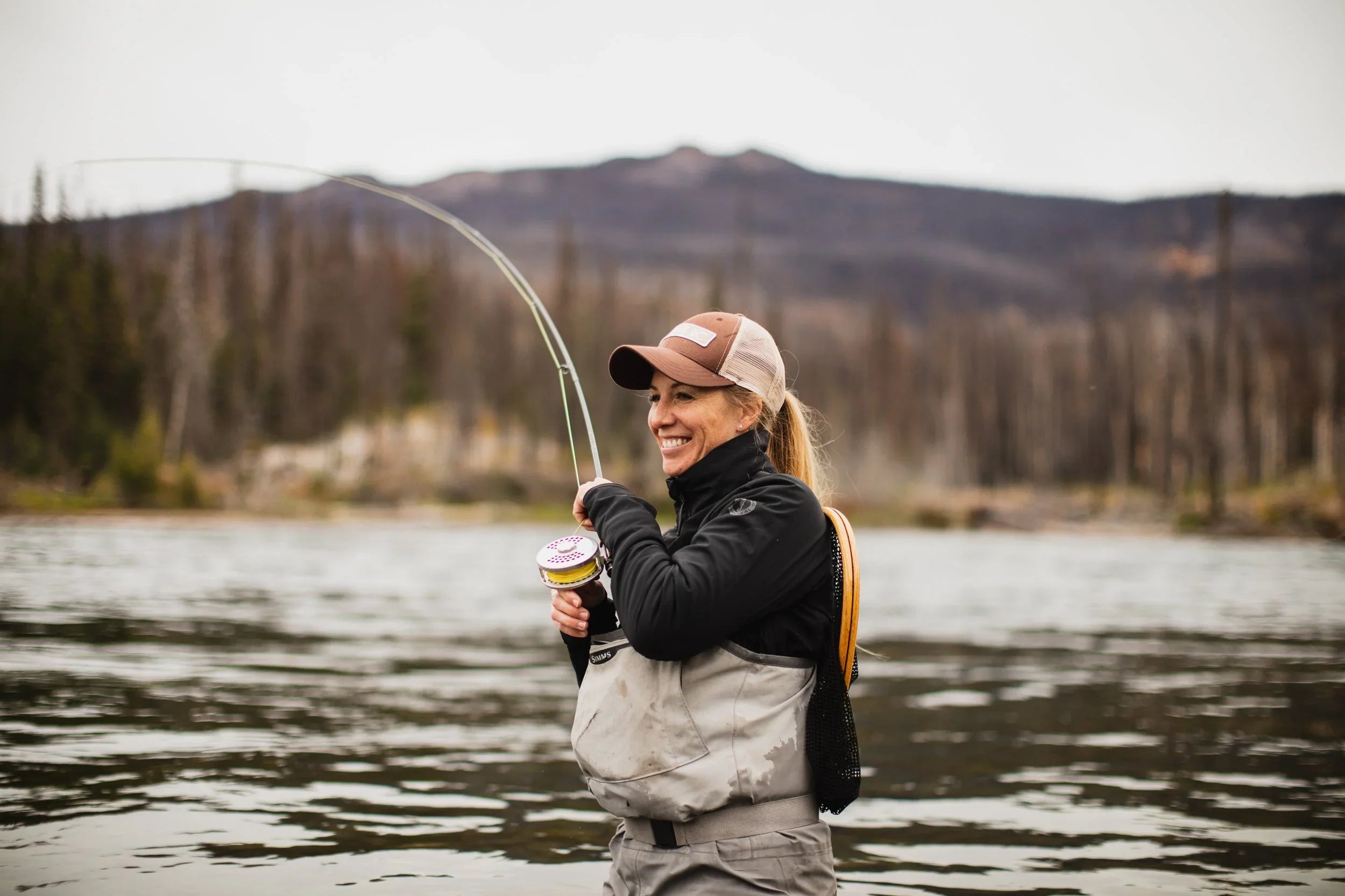 A smiling woman fly fishing while wading in a river, wearing a brown trucker hat, black jacket, and waterproof chest waders with a net tucked into her back.
