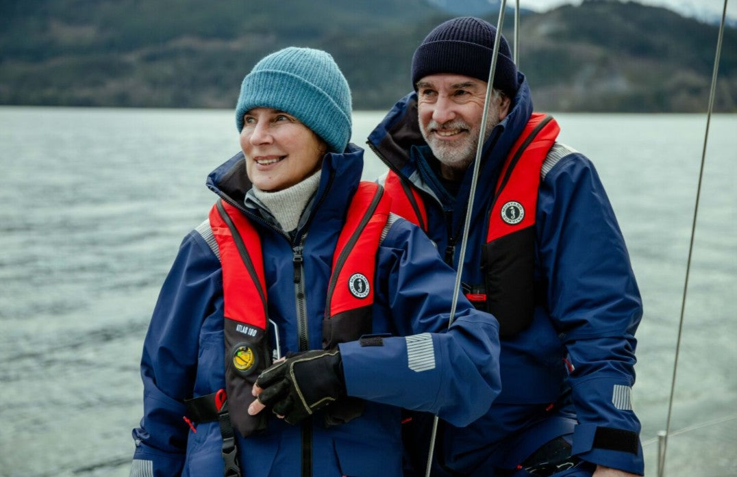 A smiling couple enjoying a cold day on the water while wearing bestselling Mustang Survival gear, including blue Meris waterproof jackets and red Atlas inflatable life jackets.