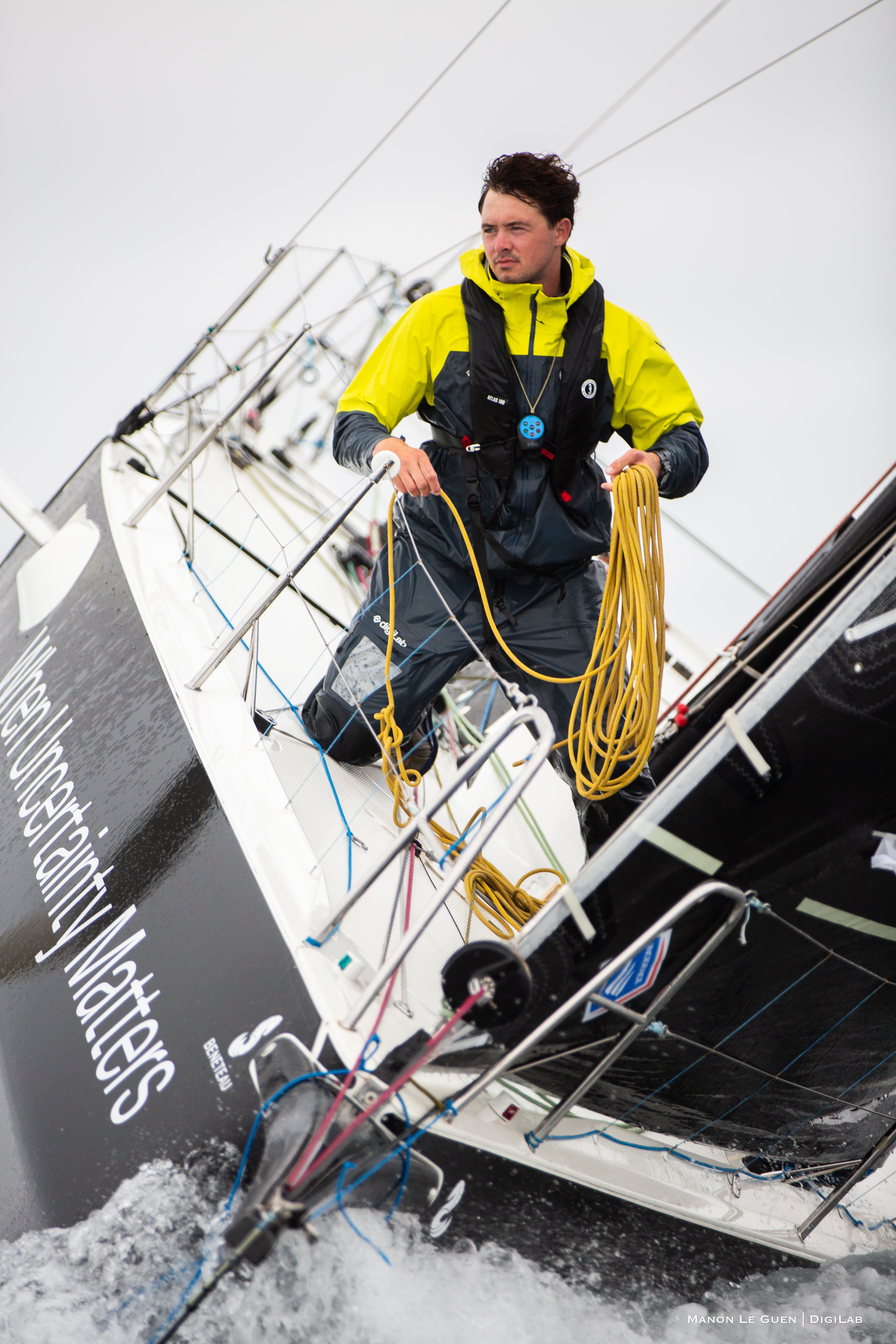 Person standing on the deck of a sailboat wearing a bright yellow and black Mustang Survival waterproof jacket and holding coiled rope, preparing lines during a sailing trip in overcast weather — great gift idea for boating or sailing enthusiasts.