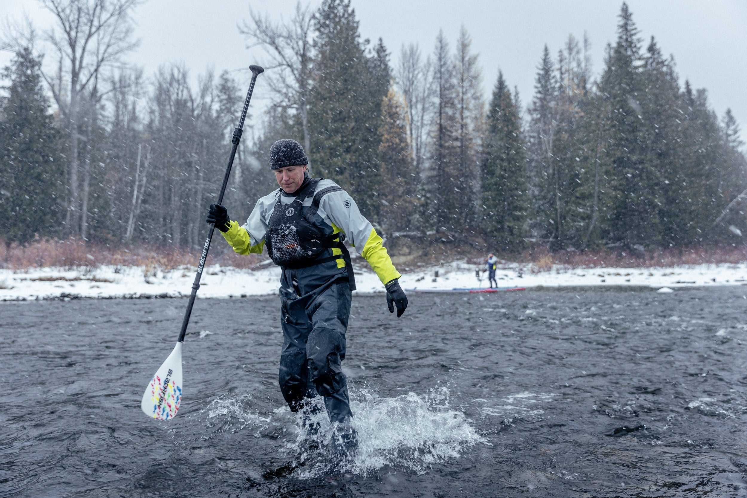 A male paddleboarder walks through a shallow river wearing a grey and high-visibility yellow Mustang Survival drysuit and a foam PFD.