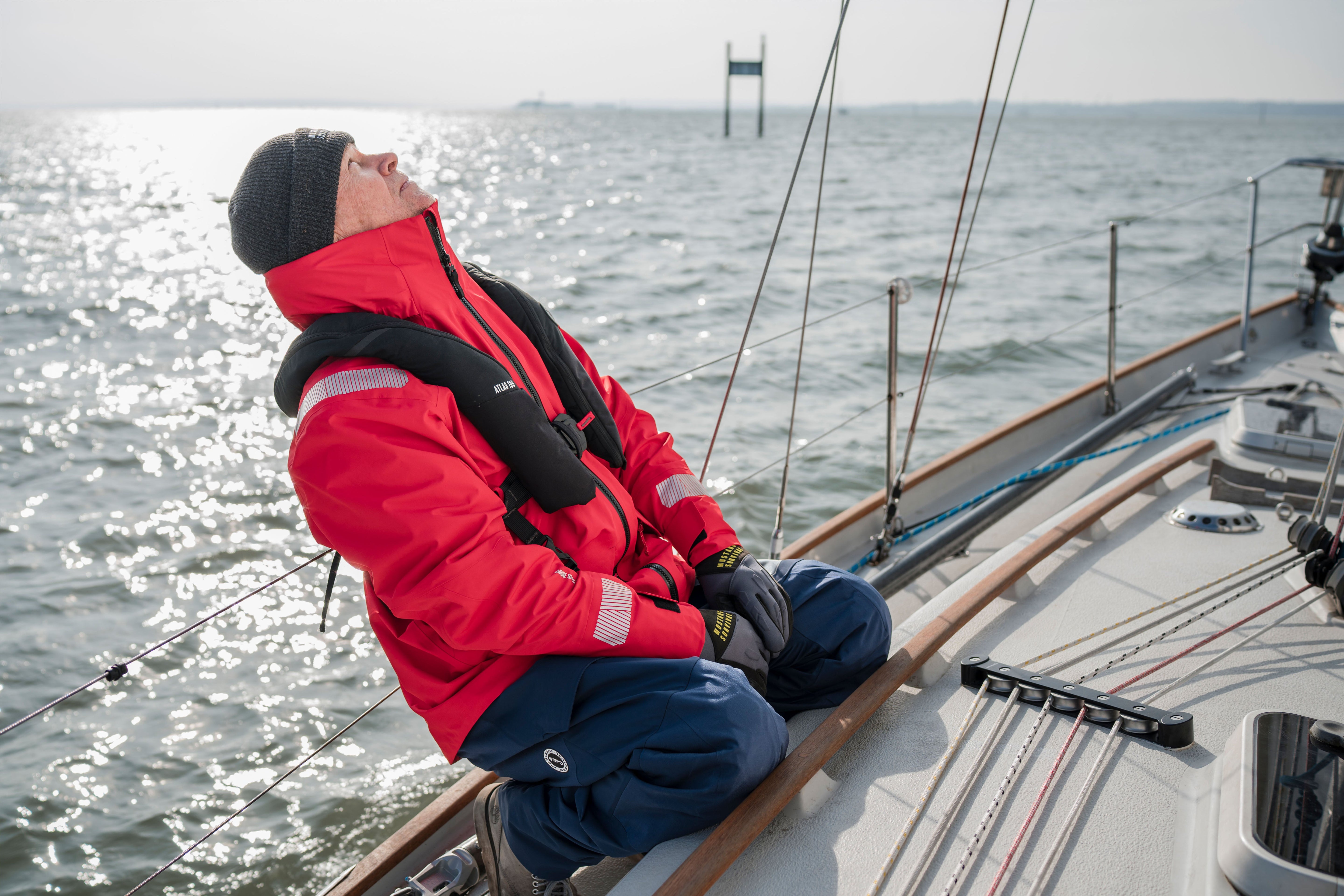 A sailor sitting on the deck of a sailboat looking upward while wearing a black beanie, a red Meris waterproof jacket, blue Meris bib pants, and a black Atlas inflatable life jacket.